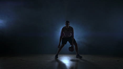 Dribbelnder-Basketballspieler-Auf-Dem-Spielfeld-Mit-Dem-Ball-In-Einem-Dunklen-Raum-Mit-Hintergrundbeleuchtung-In-Zeitlupe-Im-Rauch